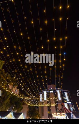 Hängende goldene Lichterketten erleuchten den festlichen Himmel einer Weihnachtsnacht mit einem modernen Gebäude und einem Weihnachtskarussell als Kulisse. Stockfoto