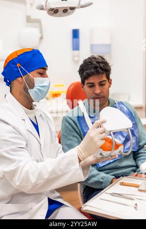 Zahnarzt mit chirurgischer Maske, Kappe und Handschuhen, die Behandlung erklären und dem Patienten in einer modernen Zahnklinik ein anatomisches Modell der Zähne zeigen, Stockfoto