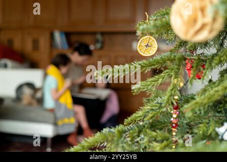 Hausgemachte Dekoration aus getrockneter Mandarine oder Orange hängt am Weihnachtsbaum. Verschwommenes Bild einer glücklichen Familie im Hintergrund Stockfoto