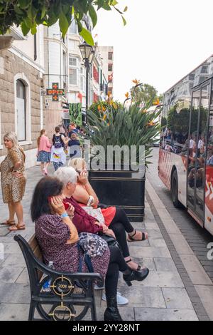 Drei Frauen sitzen auf einer Bank entlang einer belebten Straße neben einem großen Pflanzgefäß mit blühenden orangefarbenen Blumen. Gibraltar, 18. Oktober 2023 Stockfoto