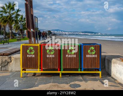 Vier Mülleimer stehen an einem Strand, jeder mit einem unterschiedlich farbigen Deckel und einem Schild mit der Aufschrift „Papier“, „Glas“, „Kunststoff“ und „organisch“. Der Strand Stockfoto