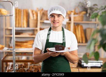 Lächelnder junger Bäcker, der in der Bäckerei Schokoladenkuchen anbietet Stockfoto