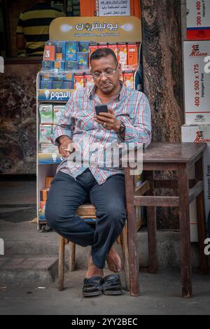 Ein Mann in Alexandria, Ägypten, sitzt vor einem Geschäft und verkauft Glühbirnen und Elektrizität mit seinem Handy. Entspannte Straßenszene des Alltags. Stockfoto