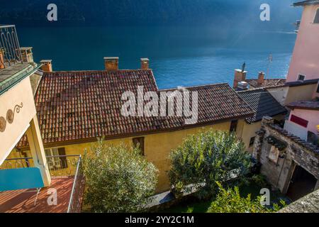 Atemberaubender sonniger Blick auf den See Lugano und die Ziegeldächer des Dorfes Gandria - Vorort der Stadt Lugano. Eine andere Küste des Sees. Rauch aus dem Kamin auf dem Dach Stockfoto