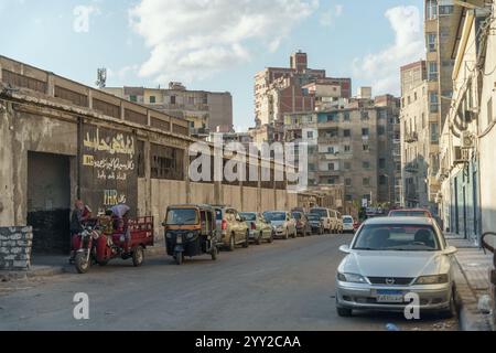 Urbane Straßenszene in Alexandria, Ägypten, mit geparkten Autos, Tuk-Tuks und alten Industriegebäuden unter klarem Himmel. Eine Mischung aus Alltag und Verfall. Stockfoto