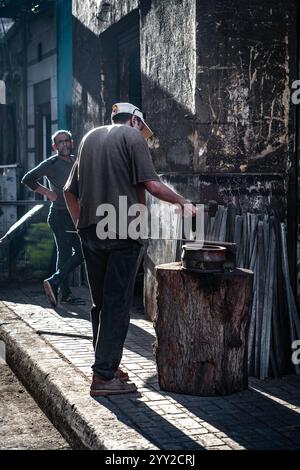 Traditioneller Schmied in Alexandria, Ägypten. Ein Mann hämmert Metall auf einen Amboss auf einem Baumstumpf, umgeben von Werkzeugen und von Sonnenlicht verfleckten Wänden Stockfoto