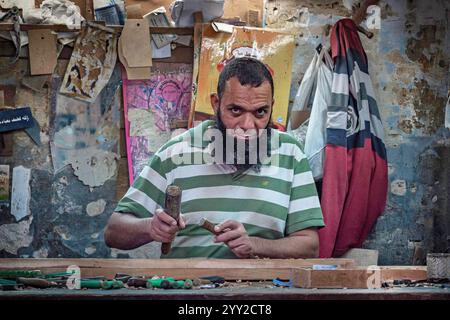 Ein Handwerker in einem gestreiften grünen Hemd arbeitet intensiv mit Werkzeugen in einer Werkstatt, die mit handgefertigtem Holz und rustikalen Wänden in Alexandria, Ägypten, gefüllt ist. Stockfoto