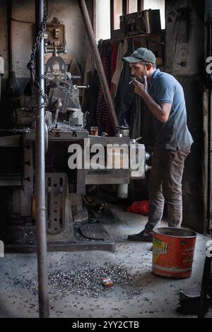 Ein Mann in einer Werkstatt, der eine Kappe trägt und neben einer Industriemaschine raucht. Der Raum ist mit Werkzeugen und Metallspänen in Alexandria, Ägypten, gefüllt. Stockfoto