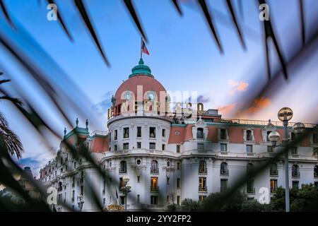 Rosafarbene Kuppel und verzierte Fassade des Negresco Hotels, einem renommierten Luxushotel in Nizza, Frankreich, das bei Sonnenuntergang durch Palmblätter vor einem farbenfrohen Himmel sichtbar ist Stockfoto