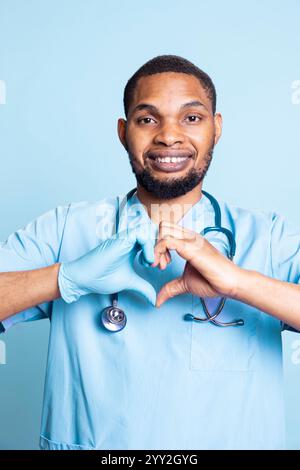 Porträt des fröhlichen Gesundheitsfachs, der Herzform-Symbol mit Gummihandschuhen an den Händen tut. Lizenzierte Krankenschwester, die im Dienst Liebesgeste zeigt, steht vor blauem Hintergrund. Stockfoto