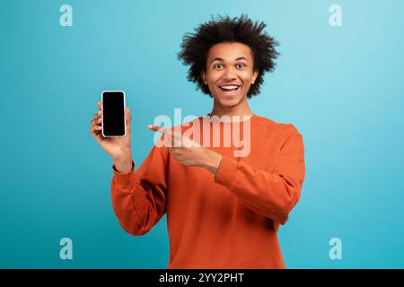 Der junge Mann mit lockigen Haaren lächelt und zeigt gegen Blau auf das Smartphone Stockfoto
