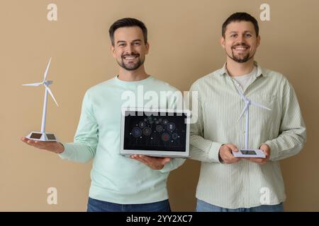 Junge Männer mit Windturbinenmodellen und Laptop auf beigefarbenem Hintergrund. Konzept der grünen Energie Stockfoto