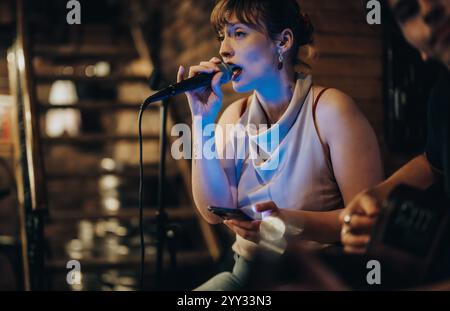 Junge Frau, die leidenschaftlich in ein Mikrofon in einem gemütlichen, intimen Musiklokal singt, umgeben von warmer Beleuchtung und lebhaftem Ambiente, um die festzuhalten Stockfoto