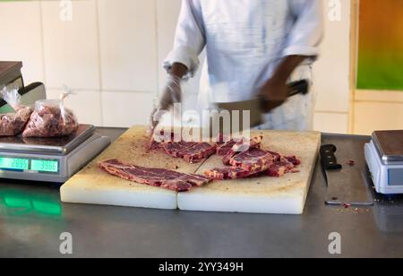 afrikanischer Metzger, der Fleischscheiben auf der Theke schneidet, Waage, die gefrorenes Rindfleisch wiegt, afroamerikaner mit einem Messer-Metzger-Beil, Stockfoto