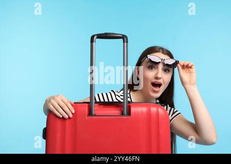 Emotionale Frau mit Koffer und Sonnenbrille auf hellblauem Hintergrund Stockfoto