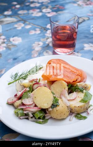 Foto einer weißen Platte mit Kartoffelsalat, Räucherlachs und Dill auf dem Teller, mit rosafarbenem Getränk im Glas dahinter, auf einer Tischdecke mit p Stockfoto