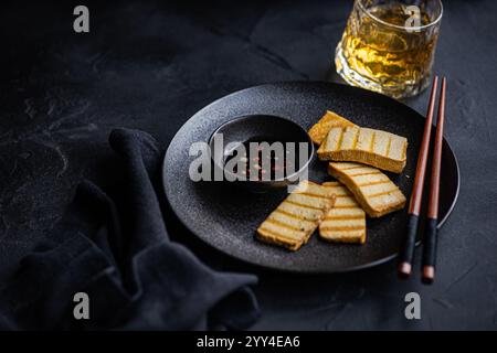 Von oben gegrillte Tofu-Soja-Käsescheiben, serviert auf einem eleganten schwarzen Teller mit würziger Pfeffersoße, begleitet von Essstäbchen und einem Glas Whiskey. Stockfoto