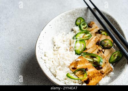 Von oben ein herzhaftes Gericht mit vom Grill gekochten Tofu-Scheiben und weißem Reis garniert mit feurig grünen Chili-Scheiben, serviert in einer gesprenkelten Schüssel mit bl Stockfoto
