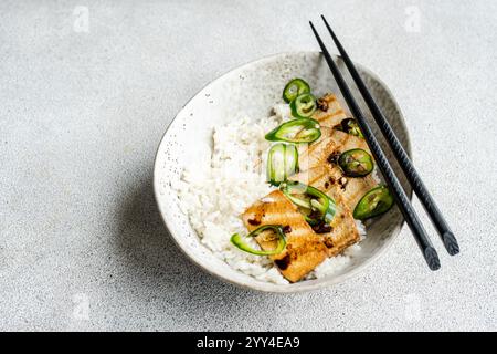 Von oben gekochte Tofu-Scheiben mit weißem Reis und scharfer grüner Paprika, begleitet von schwarzen Essstäbchen auf grauem Hintergrund. Stockfoto