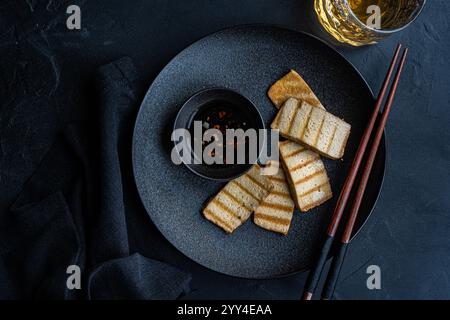 Blick von oben auf gegrillte Tofu-Scheiben, begleitet von scharfer Pfeffersoße und Essstäbchen, künstlerisch auf einem dunklen Teller mit einem Glas Getränk bes präsentiert Stockfoto