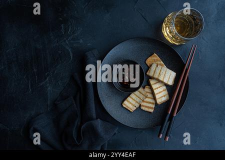 Blick von oben auf gegrillte Tofu-Soja-Käsescheiben, serviert auf einem schwarzen Teller mit Reis und scharfer Pfeffersoße, begleitet von Essstäbchen und einem Glas Getränk Stockfoto