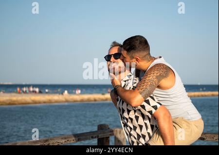 Das junge schwule Paar teilt einen freudigen Moment am Strand, verkörpert den Geist der Liebe und des Stolzes tätowierte Mann gibt einen süßen Kuss, vor einem ruhigen o Stockfoto