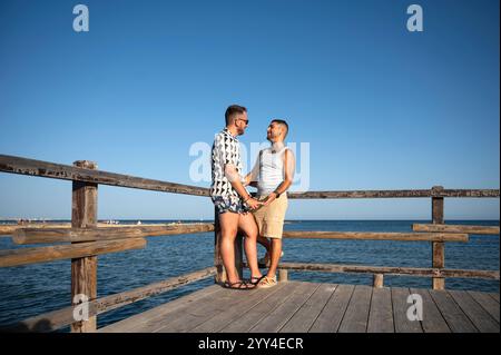 Ein junges schwules Paar genießt einen romantischen Moment am Strand und hält Hände an einem hölzernen Pier. Der klare blaue Himmel und das Meer bilden eine ruhige Kulisse für die Stockfoto