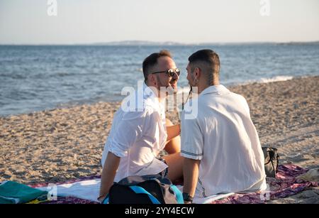 Ein junges schwules Paar genießt einen sonnigen Tag am Strand, sitzt auf einer Decke und teilt einen freudigen Moment, hervorgehoben vom Hintergrund des Ozeans und gelassen Stockfoto