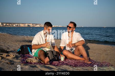 Ein junges schwules Paar genießt einen sonnigen Tag am Strand, sitzt auf einer pulsierenden Decke mit Drinks, sie sind entspannt, lächeln und verkörpern ein unbeschwertes Leben Stockfoto