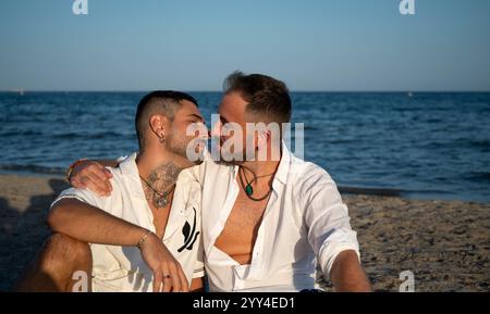 Ein schwules Paar sitzt intim am Strand und genießt einen Moment am Meer. Das Bild fängt Liebe, Stolz und Verbundenheit ein und zeigt eine wunderschöne LGBTQIA Stockfoto