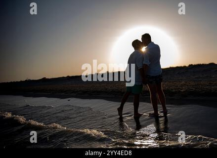 Ein junges schwules Paar genießt einen ruhigen Spaziergang am Strand bei Sonnenuntergang, symbolisiert Liebe und Freiheit, die Hände halten, sie reflektieren Glück und den Geist von p Stockfoto