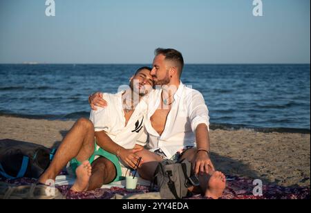 Ein junges schwules Paar genießt einen ruhigen Moment am Strand und zeigt Liebe und Intimität. Es ist entspannt auf einer Decke und genießt die Schönheit des küstenraumes Stockfoto