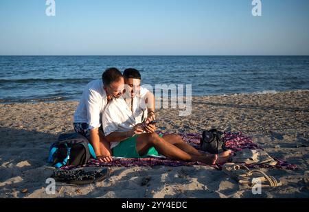 Ein fröhliches schwules Paar genießt einen sonnigen Tag am Strand, sitzt dicht auf einer Decke, die sie umarmen, während sie einen Moment mit Liebe, Entspannung und einem teilen Stockfoto