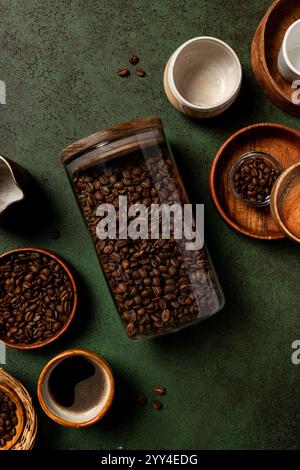 Eine gemütliche Szene mit Kaffeebohnen in einem Glasgefäß, umgeben von Keramikbechern und Holzschalen auf einer strukturierten grünen Oberfläche, perfekt für ein rustikales Kaffeematerial Stockfoto