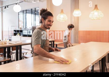 Zwei Kaffeearbeiter räumen die Tische in einem modernen Café ab. Die helle, gemütliche Inneneinrichtung mit Hängeleuchten und stilvollem Dekor sorgt für eine einladende Atmosphäre Stockfoto