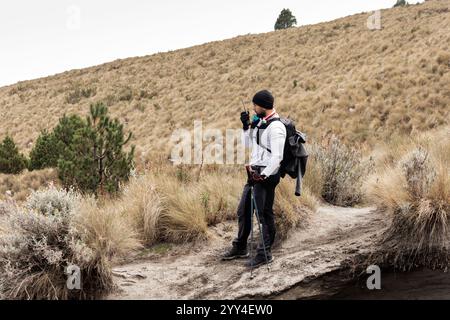 Ein schwarzer Mann in Bergsteigerausrüstung, ausgestattet mit Rucksack und Wanderstock, steht auf einem schroffen Weg. Er kommuniziert über Walkie-Talkie zwischen einer Halterung Stockfoto