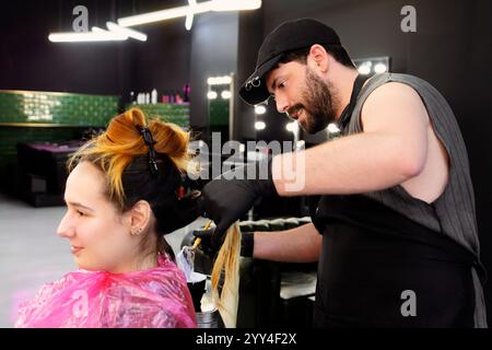 Ein erfahrener Stylist mit Handschuhen trägt Haarfarbe auf das Haar eines Kunden in einem eleganten, gut beleuchteten Salon auf. Die Atmosphäre ist modern und professionell, betont Stockfoto