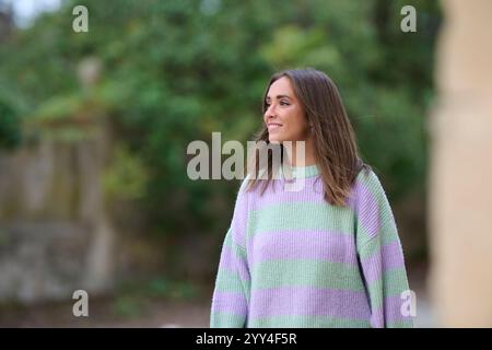 Eine junge Frau in einem Pastellstreifen-Pullover steht draußen und lächelt. Der sanfte Fokus auf dem Hintergrund unterstreicht die natürliche Umgebung und bietet eine se an Stockfoto