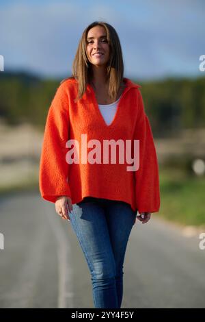 Eine Frau in einem orangefarbenen Pullover und einer Jeans geht selbstbewusst auf einer sonnigen Straße, die sich im natürlichen Licht sonnt. Der malerische Hintergrund verleiht dem VI eine ruhige Note Stockfoto