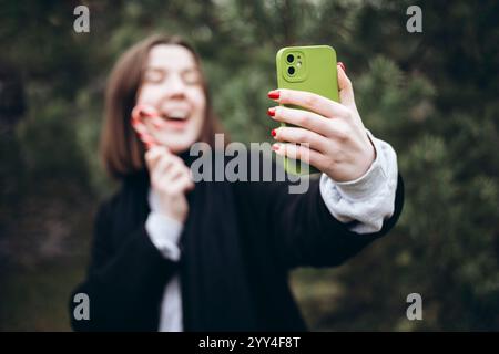 Eine fröhliche Person macht ein Selfie mit einem Zuckerrohr im Freien, das den festlichen Geist verkörpert. Die grüne Handyhülle steht im Kontrast zu roten Nägeln, was die noch ergänzt Stockfoto