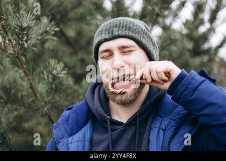 Ein bärtiger Mann in einer Winterjacke und Strickmütze genießt einen Zuckerrohr im Freien, umgeben von immergrünen Bäumen, er umrahmt den festlichen Geist des Urlaubs Stockfoto
