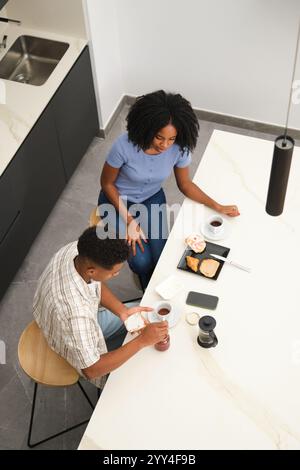 Ein junges afroamerikanisches Paar genießt ein gemütliches Frühstück in ihrer modernen Küche, sie teilen sich Kaffee und Gebäck und schaffen eine warme Atmosphäre voller Zuhause Stockfoto