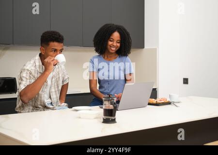 Ein junges schwarzes Paar genießt einen entspannten Morgen in ihrem modernen Zuhause, sie teilen sich Kaffee und Gebäck an der Küchentheke, während sie auf einem stöbern Stockfoto