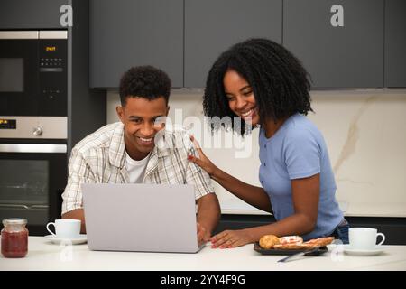 Ein junges schwarzes Paar genießt einen gemütlichen Moment zu Hause, indem es sich mit einem Laptop beschäftigt, während das Frühstück auf den Tisch gelegt wird. Es ist entspannt und lächelnd und schafft so einen Stockfoto