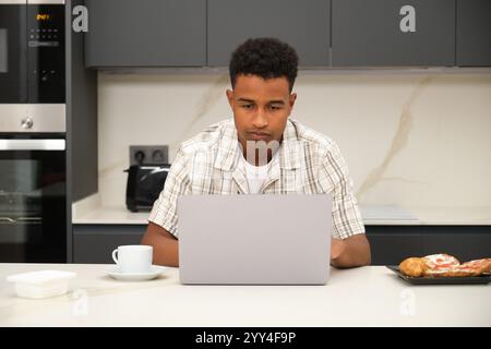 Ein junger Schwarzer sitzt bequem in seiner modernen Küche und teilt einen Laptop. Dieses Bild fängt einen Moment der Entspannung und Verbindung in einem gemütlichen Zuhause ein Stockfoto