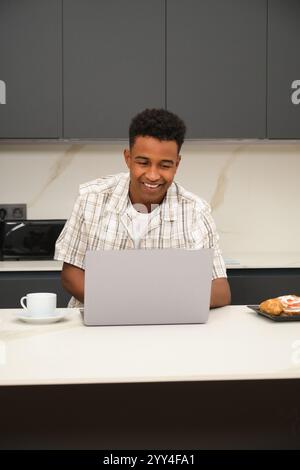 Ein fröhlicher junger schwarzer Mann verbringt gute Zeit in ihrer modernen Küche. Der Mann lächelt an seinem Laptop und zeigt eine entspannte, glückliche Umgebung Stockfoto