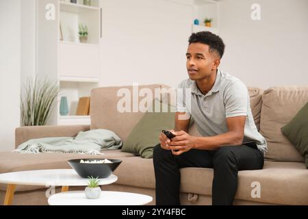 Ein junger afroamerikanischer Mann genießt einen entspannten Tag zu Hause. Er sitzt bequem auf einem beigefarbenen Sofa mit Fernbedienung in der Hand, umgeben von modernem Deko Stockfoto