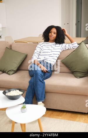 Eine junge schwarze Frau sitzt bequem auf einem beigefarbenen Sofa in einem modernen Wohnzimmer. Sie trägt ein gestreiftes Hemd und eine Jeans und sieht mit einer Schüssel Popco entspannt aus Stockfoto