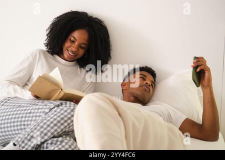 Ein junges afroamerikanisches Paar genießt einen friedlichen Tag zu Hause. Die Frau liest ein Buch, während der Mann sich mit einem Smartphone entspannt und ein gemütliches und inniges Ambiente verkörpert Stockfoto