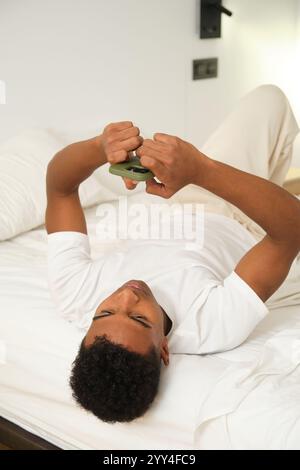 Ein junger Afroamerikaner liegt auf einem Bett in einem hellen, modernen Zimmer und hält ein Smartphone. Er trägt ein weißes Hemd und genießt Freizeit tim Stockfoto